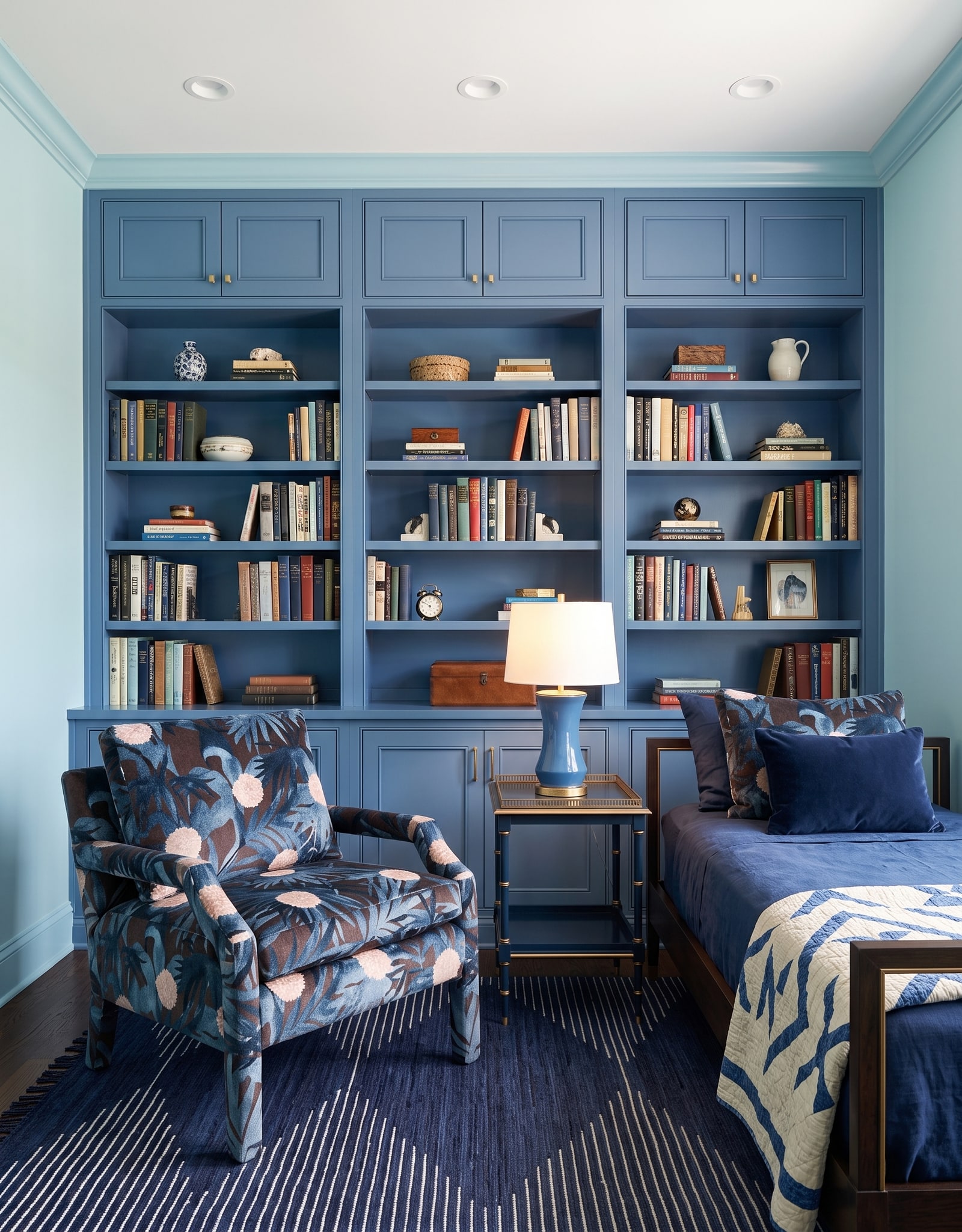 A medium-blue bedroom bookcase with supported shelves, closed cabinets above and below, a chair, side table, and bed in front.