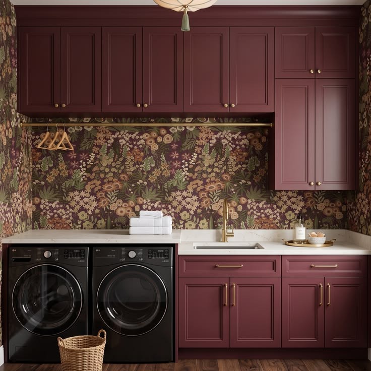 Maroon laundry cabinets surrounding side-by-side machines beneath dark floral wallpaper.