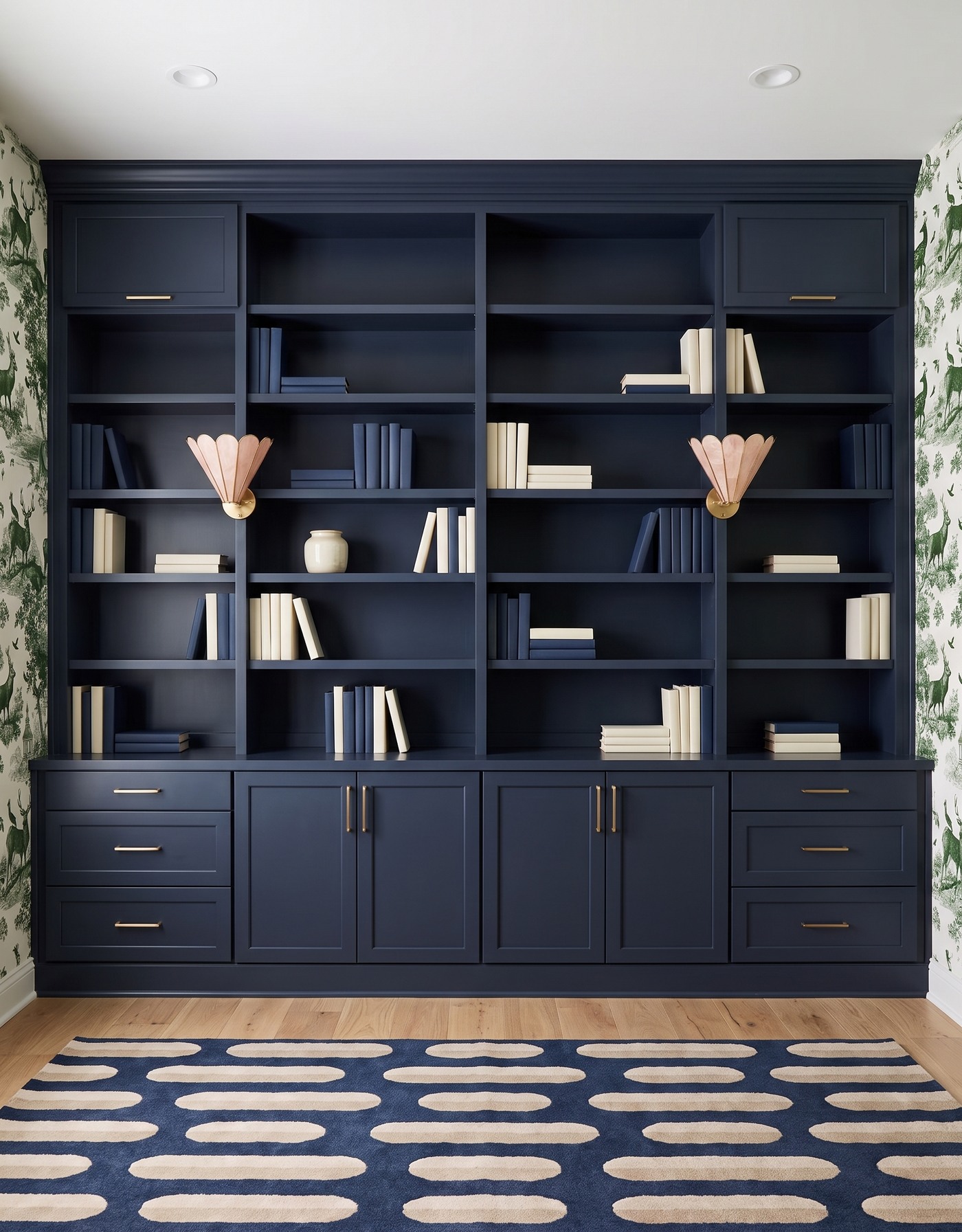 Library with navy framed cabinets and solid shelf backs, green-and-ivory landscape toile side walls, pale wood floor, navy-and-cream capsule-pattern rug, blush shell sconces and navy-and-ivory books.