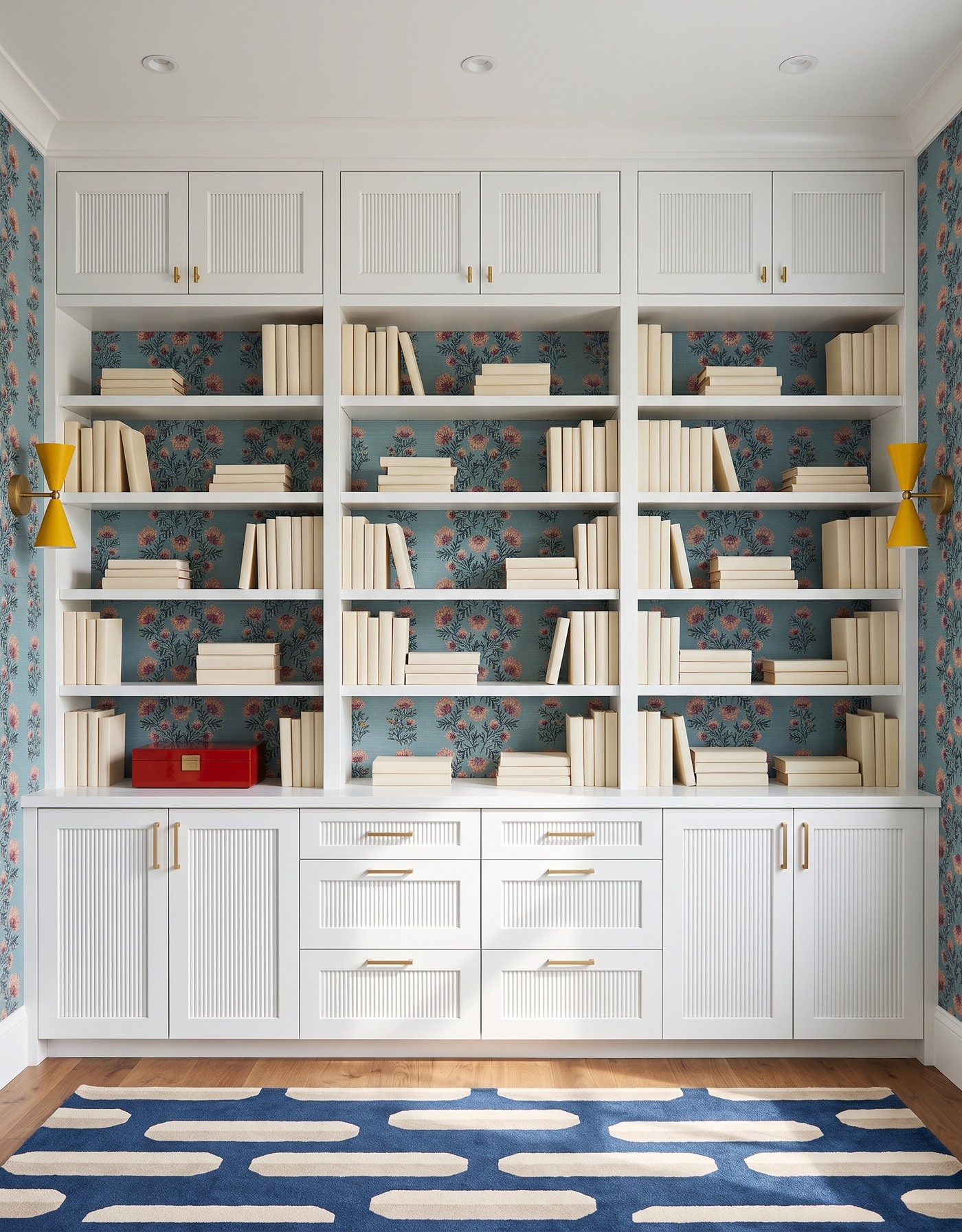 Library with white grooved cabinet fronts, dusty blue floral wallpaper with pink-and-ochre blossoms, honey wood floor, navy-and-cream capsule-pattern rug, mustard hourglass sconces and ivory books.