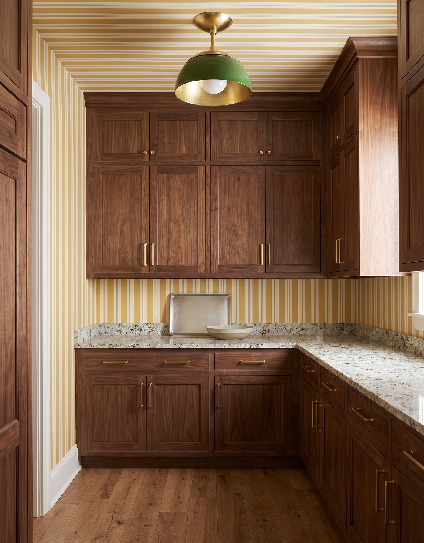 Pantry with walnut cabinets, mustard-and-ivory narrow striped wallpaper, granular cream counter, warm wood floor and green pendant.