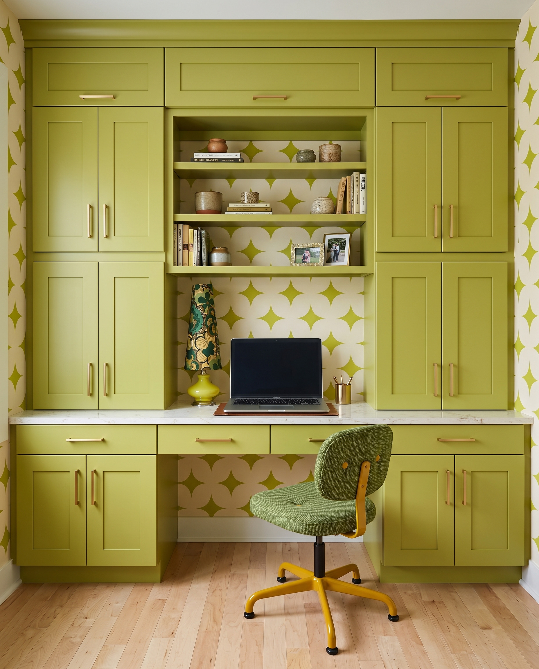 Olive-chartreuse built-in desk with tall closed cabinets, cream geometric wallpaper, open central shelves and a green swivel chair.