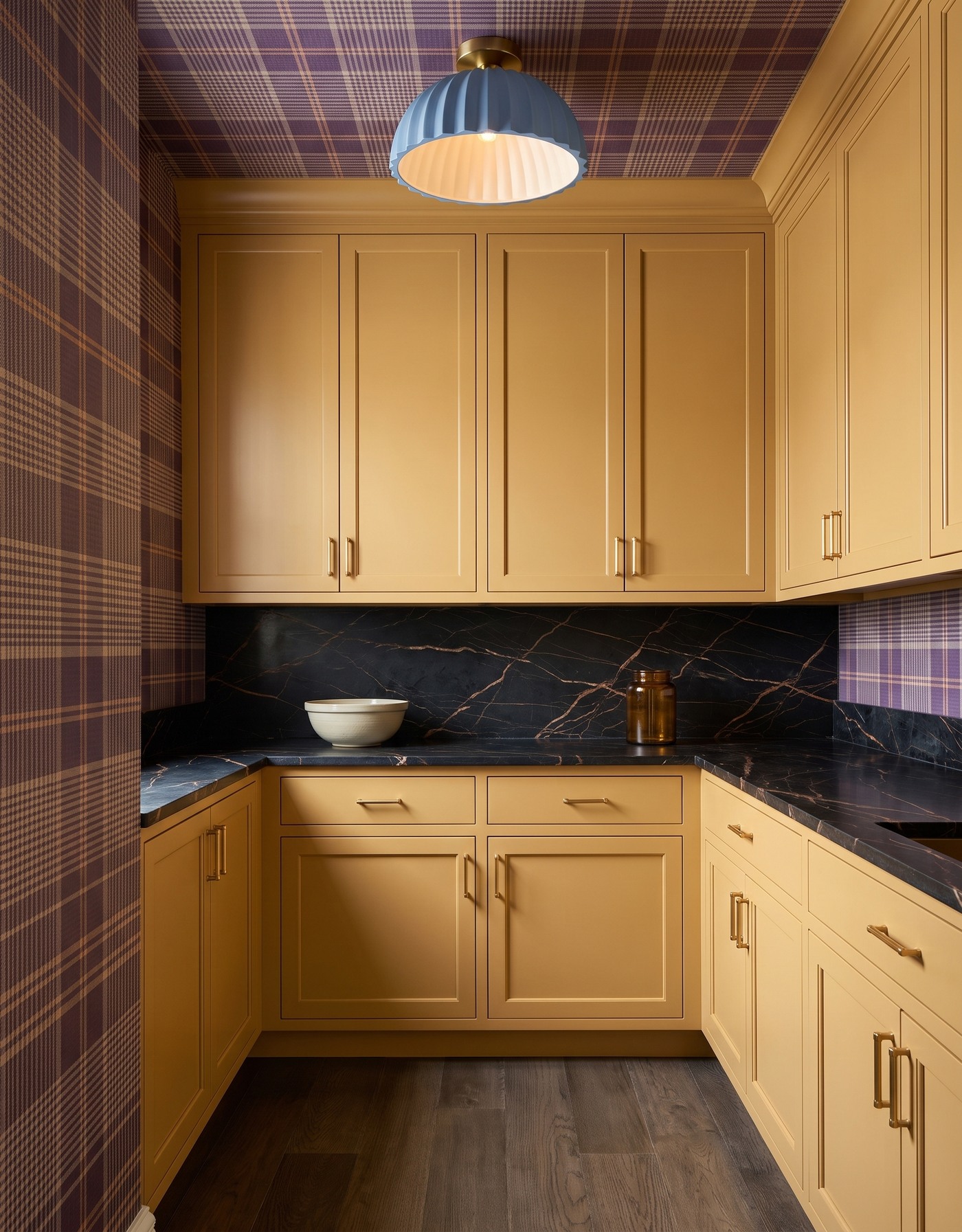 Pantry with ochre cabinets, plum-and-tan plaid wallpaper, charcoal bronze-veined counters, gray-brown wood floor and dusty blue pleated light.