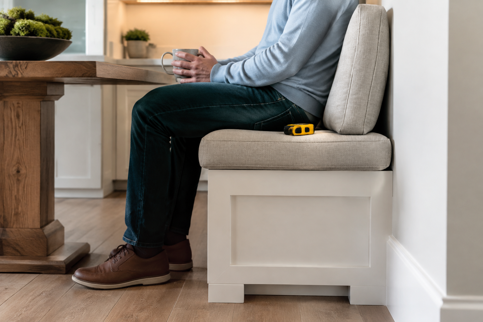 A seated adult rests naturally against an angled banquette cushion with feet flat on the floor and a tape measure on the seat.