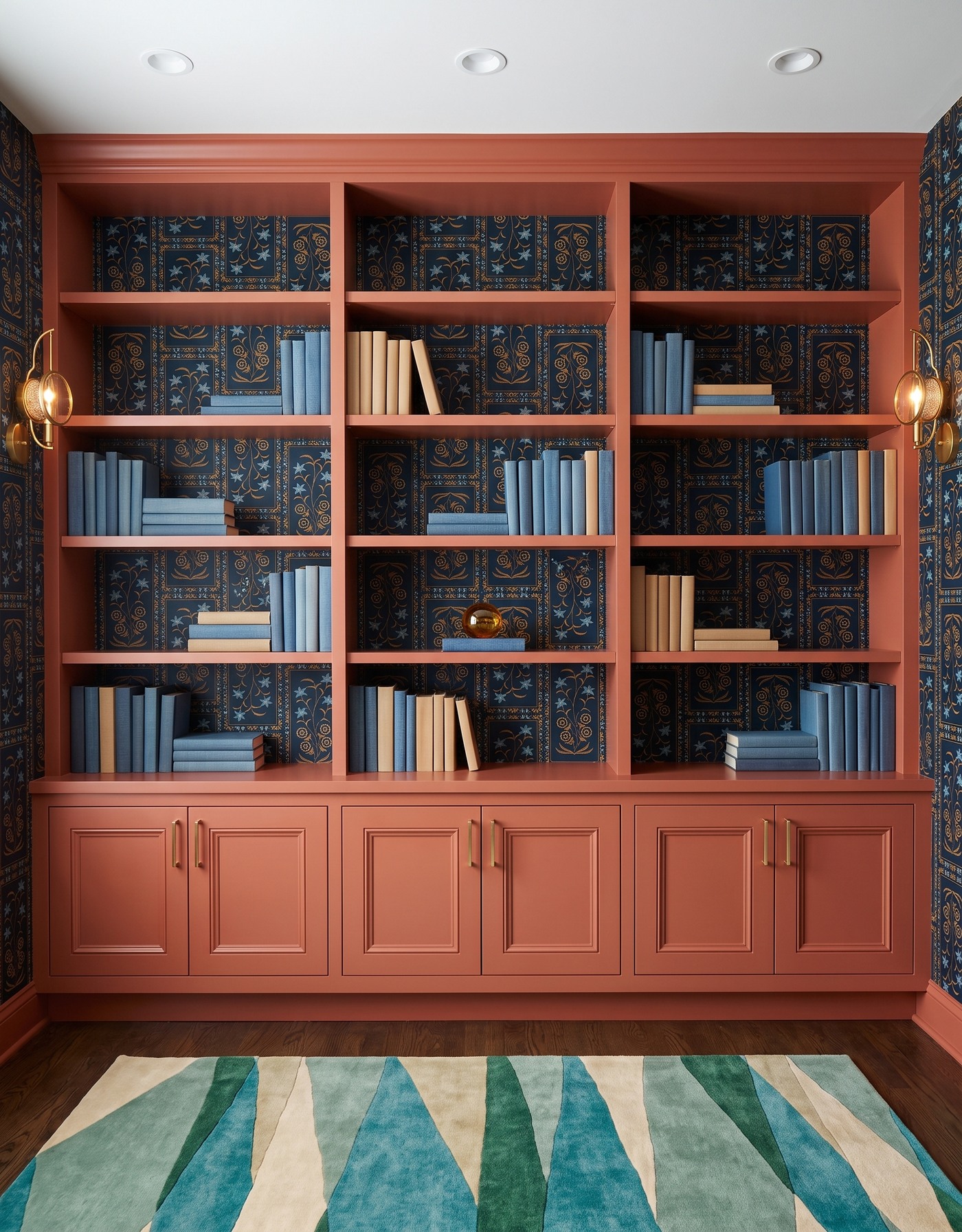 Library with rust framed base cabinets and shelving, navy-and-gold framed botanical wallpaper, brown wood floor, teal-and-cream angular rug, brass-and-amber circular sconces and blue-and-cream books.