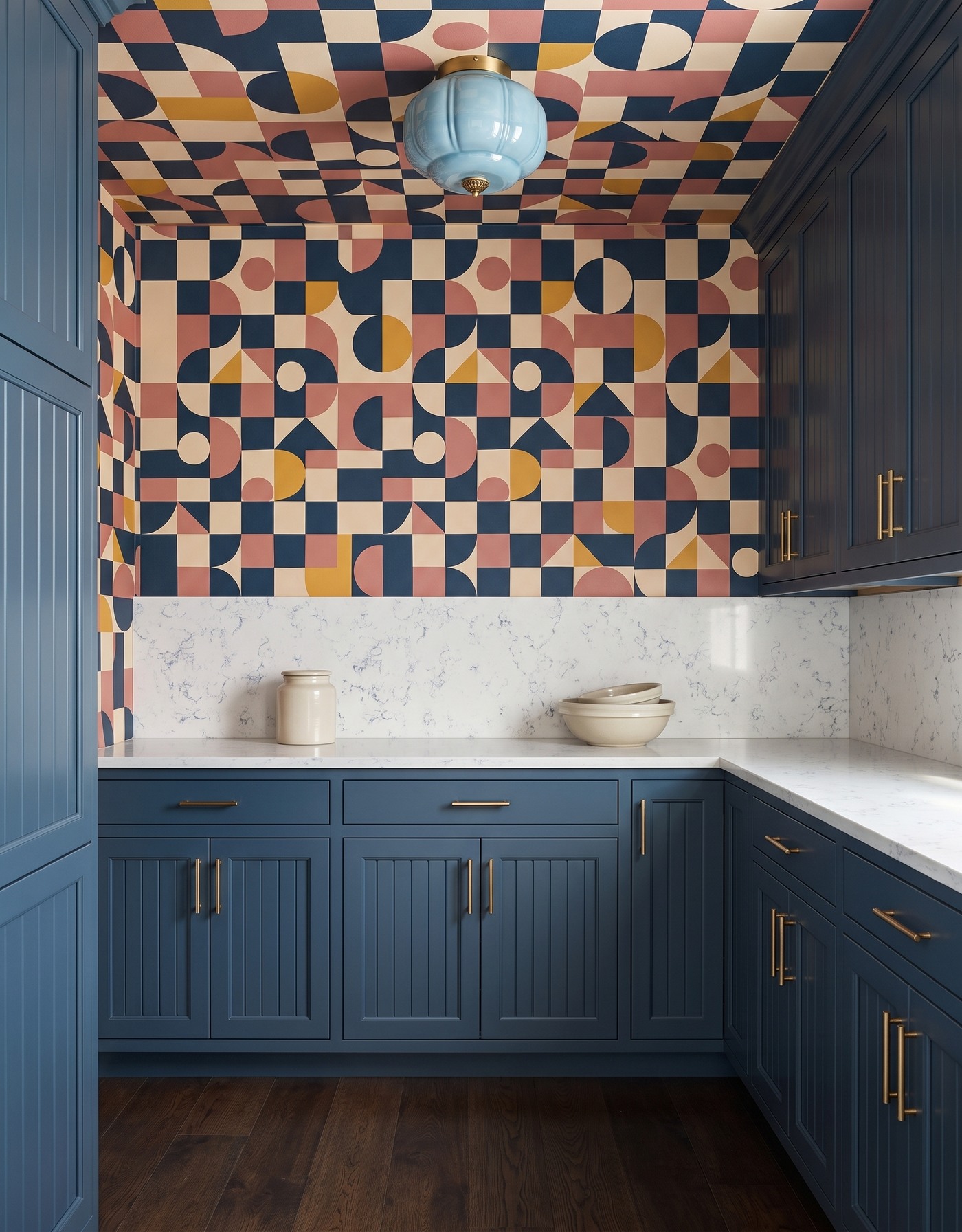 Pantry with blue vertically textured cabinets, navy-coral-mustard-and-ivory geometric wallpaper, white fine gray-veined counter, dark wood floor and blue glass light.