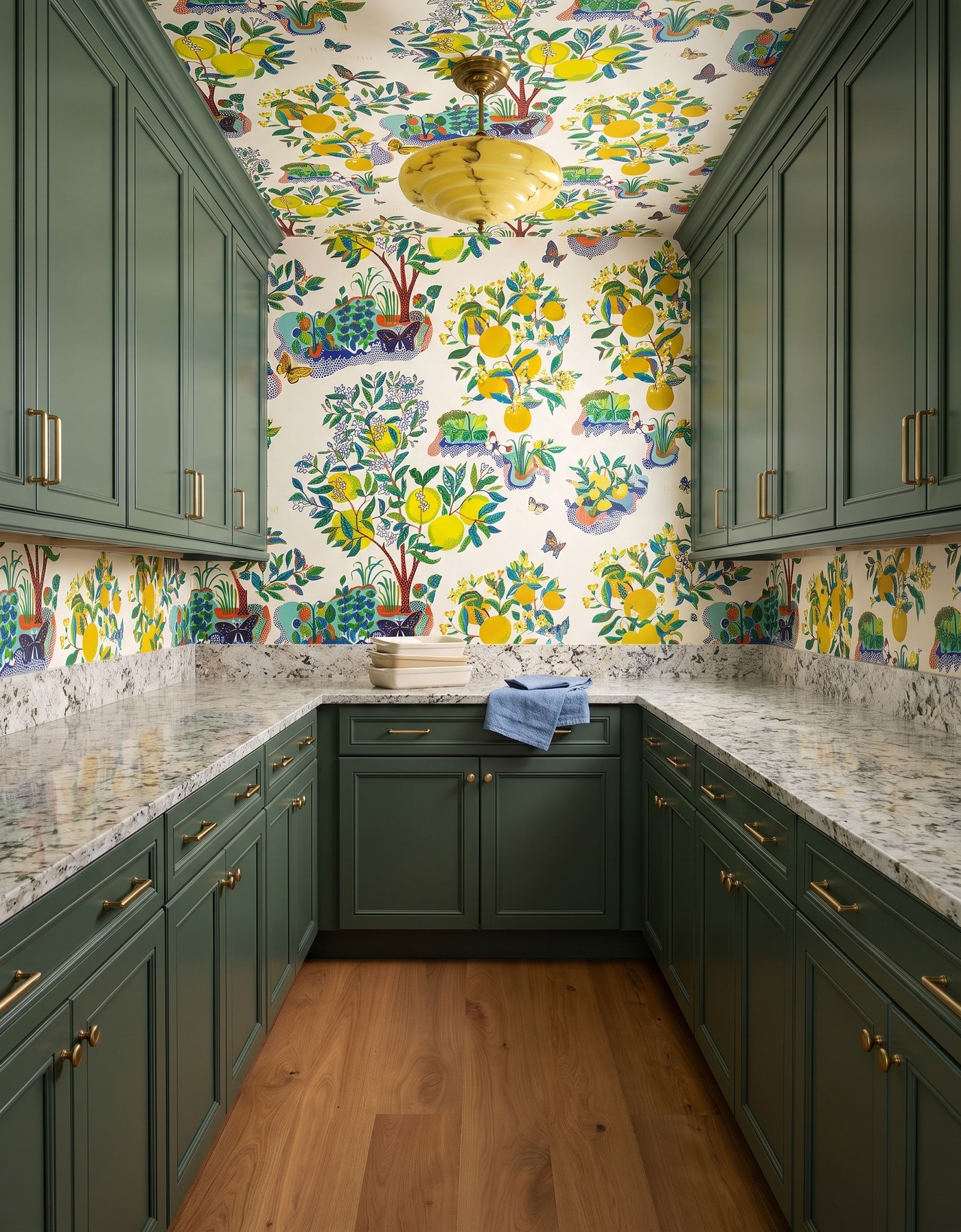 Pantry with forest green cabinets, multicolor fruit-and-botanical wallpaper, granular cream counter, warm wood floor and yellow glass light.