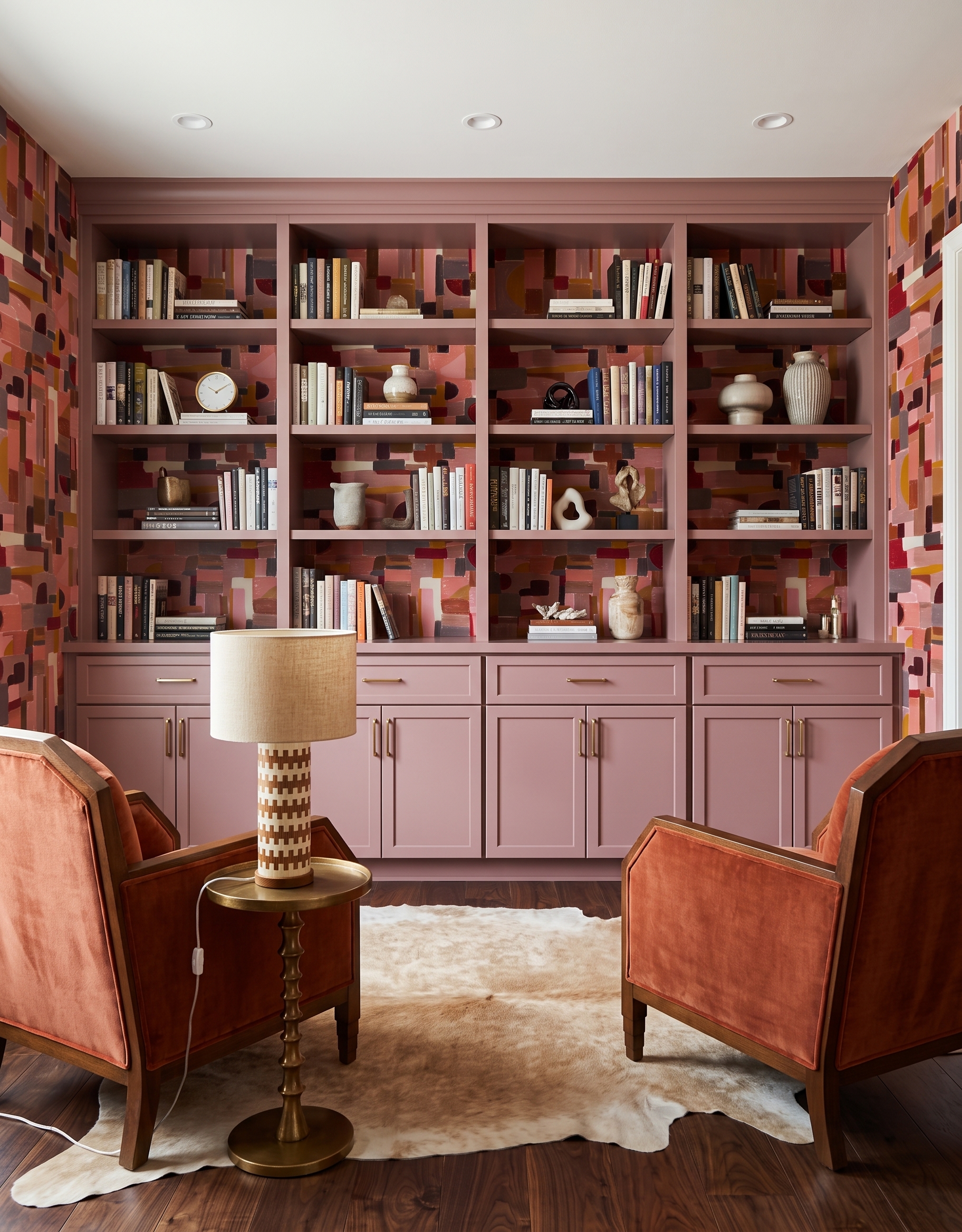 Dusty-rose bookcase concept with block-patterned wallpaper, two rust upholstered wood-frame chairs, a cream rug and a geometric table lamp.